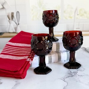 May include: Three ruby red wine glasses with ornate floral designs. The glasses are on a marble surface, with a red and white striped towel and a white kitchen background. The glasses have a decorative stem and base.