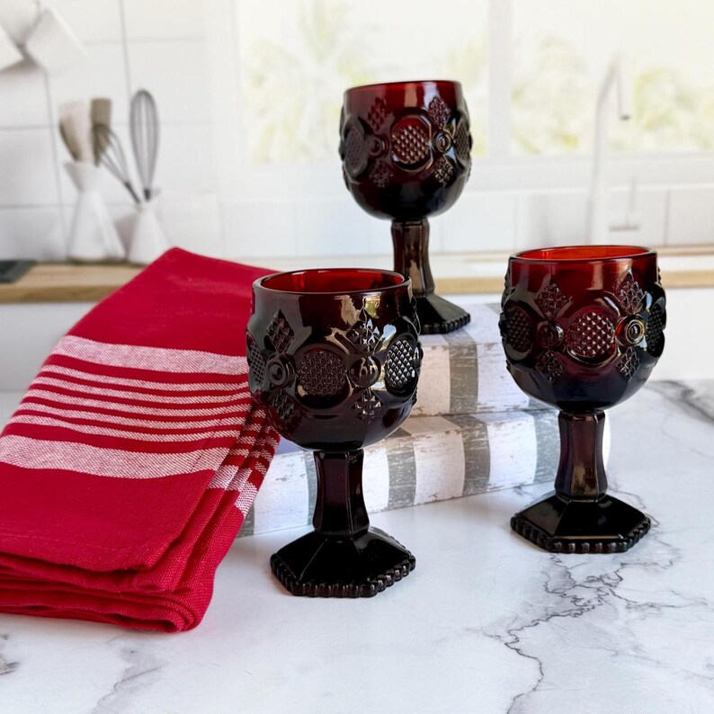 May include: Three ruby red wine glasses with ornate floral designs. The glasses are on a marble surface, with a red and white striped towel and a white kitchen background. The glasses have a decorative stem and base.