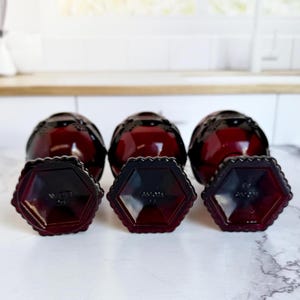 May include: A collection of three dark red glass bottles with matching hexagonal bases. The bottles have a textured, decorative design and are arranged on a white surface. The bases are also dark red and feature the word "AVON".