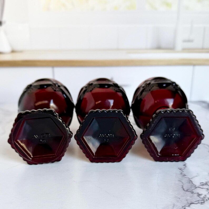 May include: A collection of three dark red glass bottles with matching hexagonal bases. The bottles have a textured, decorative design and are arranged on a white surface. The bases are also dark red and feature the word "AVON".