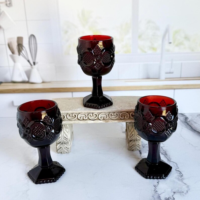 May include: Three ruby red goblets with ornate floral and geometric designs. The wine glasses have a dark stem and base, and a bright red interior. The goblets are displayed on a small, decorative table.