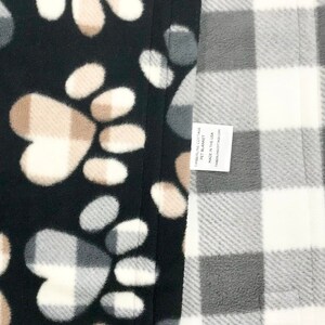 Puede incluir: Una manta para mascotas negra y blanca con un dise&ntilde;o de huella de pata en un lado y un dise&ntilde;o de cuadros grises y blancos en el otro lado. La manta tiene una etiqueta que dice "Timberline Cottage Pet Blanket Made in the USA Timberlinecottage.com".