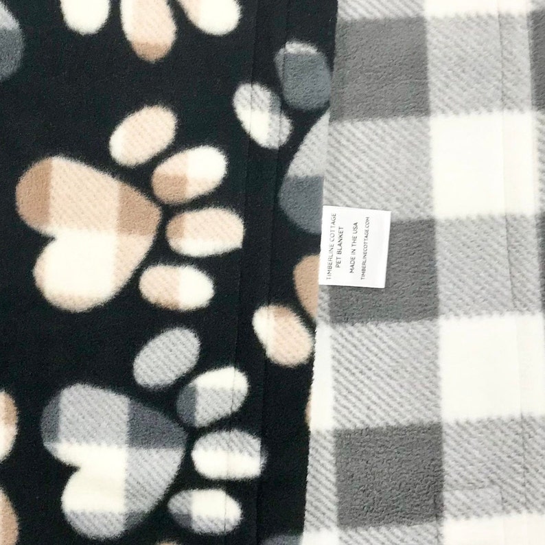 Puede incluir: Una manta para mascotas negra y blanca con un dise&ntilde;o de huella de pata en un lado y un dise&ntilde;o de cuadros grises y blancos en el otro lado. La manta tiene una etiqueta que dice "Timberline Cottage Pet Blanket Made in the USA Timberlinecottage.com".