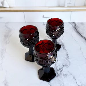 May include: Three ruby red goblets with ornate floral and geometric patterns. The goblets have a hexagonal base and a detailed stem. The glass has a rich, dark red color, and the design is repeated around the bowl of the glass.