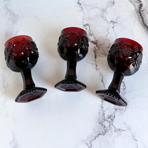 May include: Three ruby red vintage wine glasses. Each glass features a detailed pattern on the bowl and a faceted base. The glasses are arranged on a white and gray marble-like surface, showcasing their rich color and design.