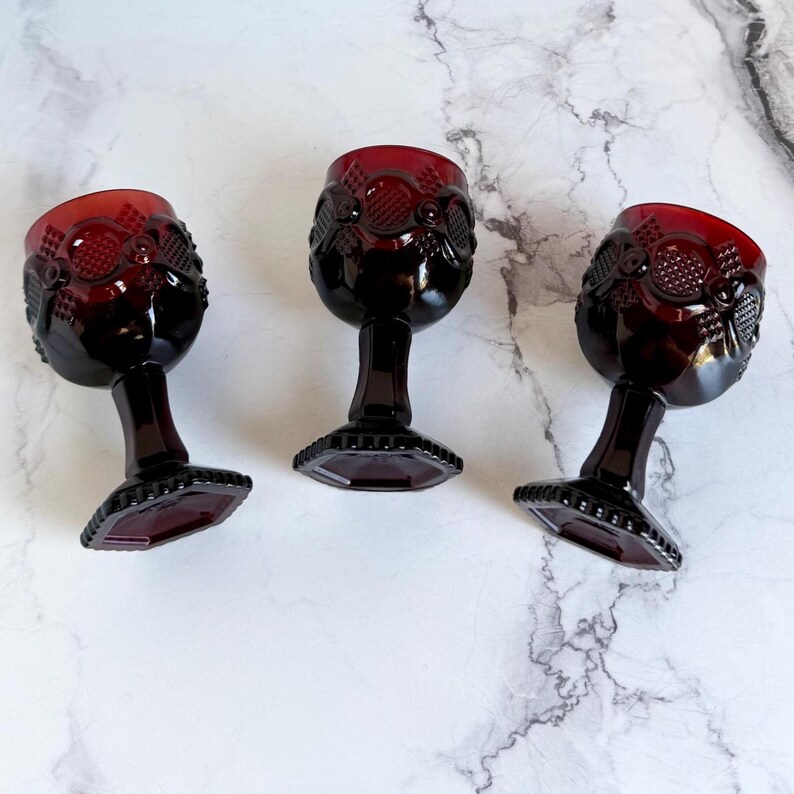May include: Three ruby red vintage wine glasses. Each glass features a detailed pattern on the bowl and a faceted base. The glasses are arranged on a white and gray marble-like surface, showcasing their rich color and design.