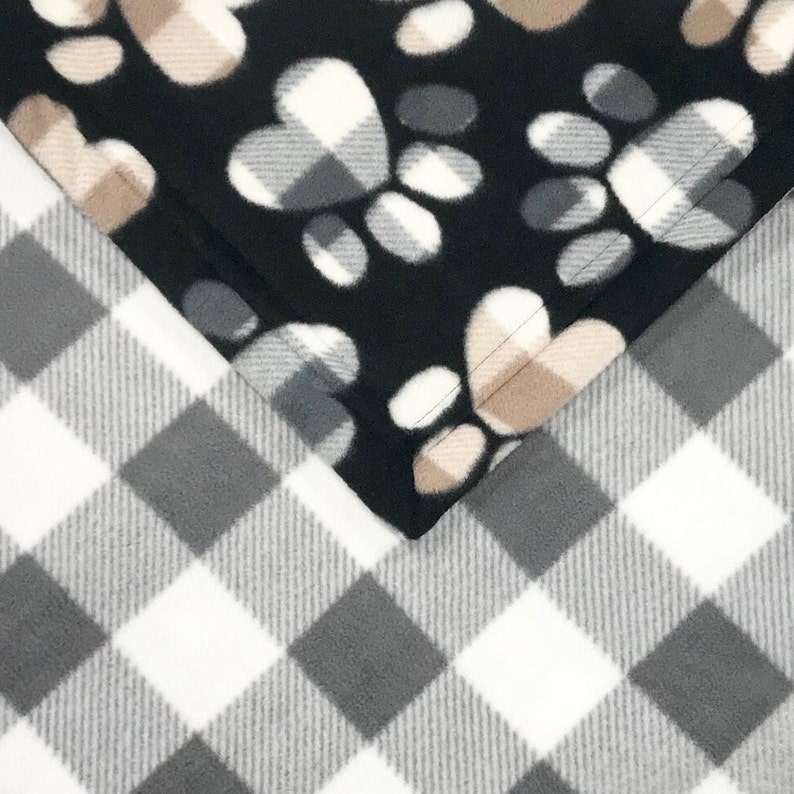 Puede incluir: Una manta de forro polar reversible con un fondo negro y un patr&oacute;n de huellas de patas en un lado y un patr&oacute;n de cuadros de b&uacute;falo blanco y gris en el otro.