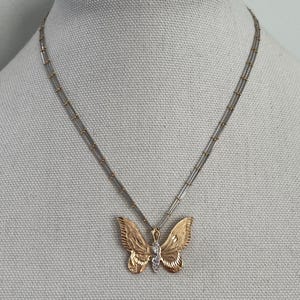 May include: A gold-coloured butterfly pendant necklace with a silver chain. The butterfly has detailed wing patterns and a small diamond-like accent in the centre. The necklace is displayed on a mannequin.