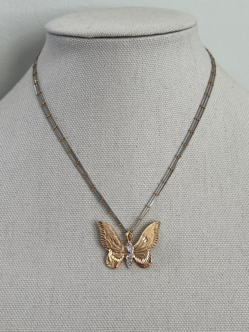 May include: A gold-coloured butterfly pendant necklace with a silver chain. The butterfly has detailed wing patterns and a small diamond-like accent in the centre. The necklace is displayed on a mannequin.