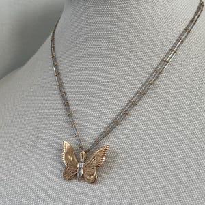 May include: A gold butterfly pendant necklace with a small diamond. The butterfly has detailed wing patterns and hangs from a delicate, multi-strand chain. The necklace is displayed on a neutral-coloured mannequin.
