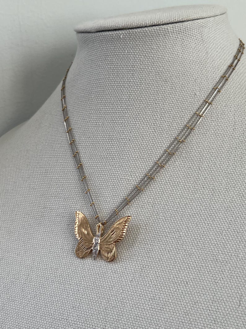 May include: A gold butterfly pendant necklace with a small diamond. The butterfly has detailed wing patterns and hangs from a delicate, multi-strand chain. The necklace is displayed on a neutral-coloured mannequin.