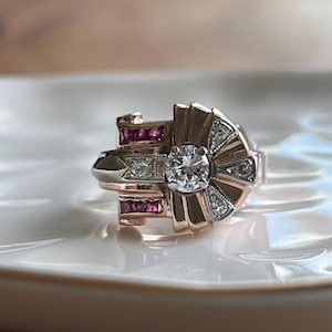 Peut inclure: Bague de style Art Déco avec un diamant rond central et de petits diamants d'appoint. La bague présente un design géométrique avec des tons or rose et argent, et de petites pierres précieuses rouges. La bague est posée sur une surface blanche.