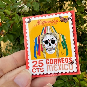 May include: A red, white, and yellow sticker with a sugar skull wearing a colorful hat and the text "Michoacán Día de los Muertos 25 Correos Cts México @comadreculture".