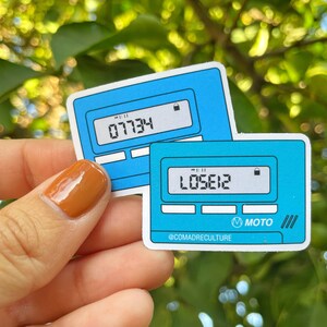 May include: Two blue and white stickers with a calculator design. The top sticker displays the number "07734" and the bottom sticker displays the number "L0562". The stickers have the text "@COMADRECULTURE" and "MOTO" on them.