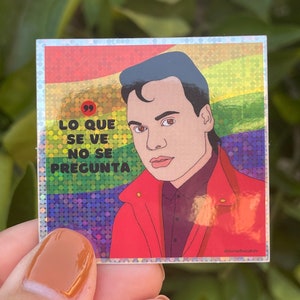 May include: A sticker with a rainbow background and a cartoon illustration of a person with a red jacket. The sticker has the text "Lo que se ve no se pregunta" in black.