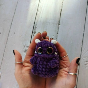 Puede incluir: Un peque&ntilde;o monstruo de crochet morado con detalles blancos y grandes ojos negros. El monstruo est&aacute; siendo sostenido en las manos de una persona.