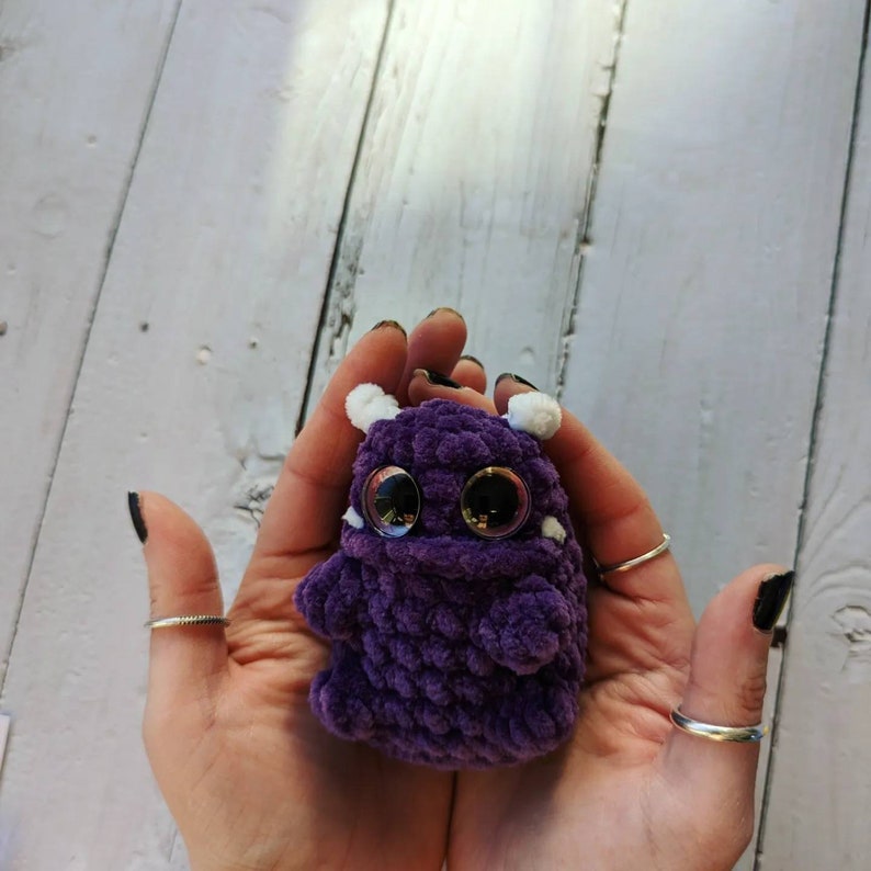 Puede incluir: Un peque&ntilde;o monstruo de crochet morado con detalles blancos y grandes ojos negros. El monstruo est&aacute; siendo sostenido en las manos de una persona.