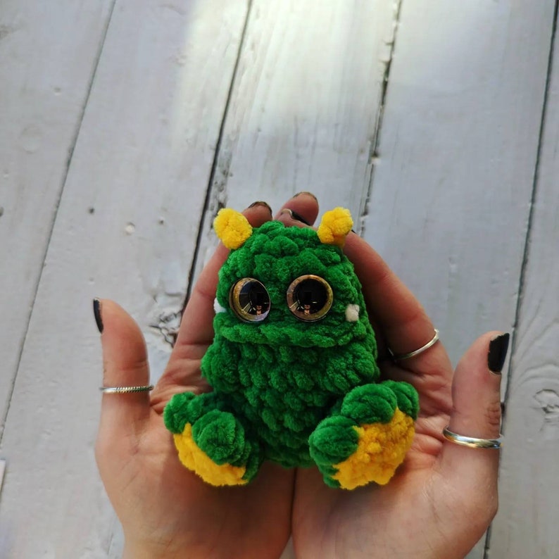 Puede incluir: Un juguete monstruo de crochet verde con detalles de hilo amarillo y grandes ojos saltones. El juguete est&aacute; siendo sostenido en las manos de una persona.