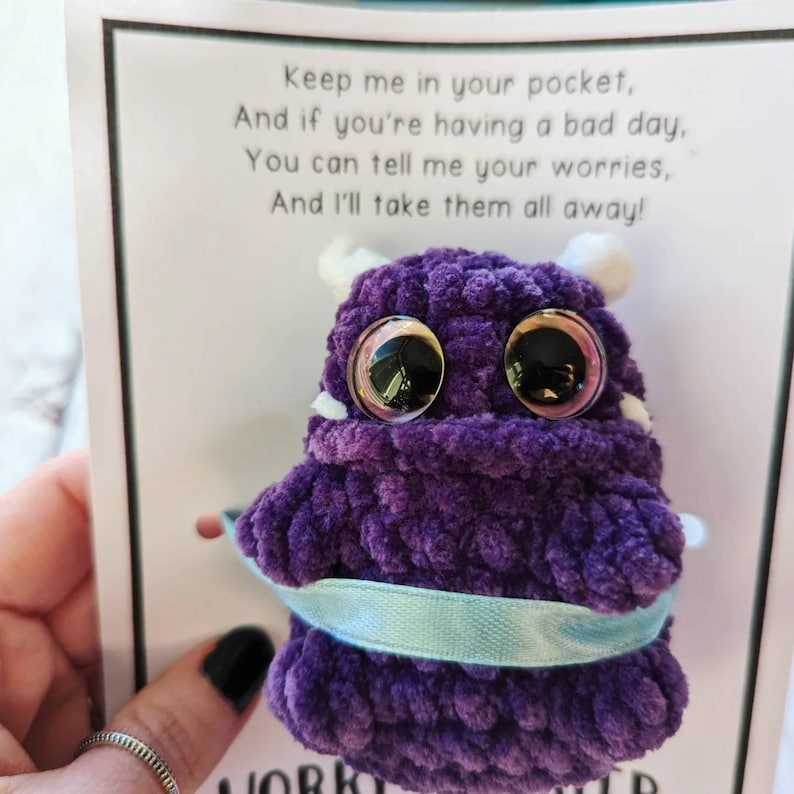 Puede incluir: Monstruo de crochet morado con grandes ojos saltones y una cinta azul claro atada alrededor. El monstruo est&aacute; sentado en una tarjeta blanca con el texto "Keep me in your pocket, And if you're having a bad day, You can tell me your worries, And I'll take them all away!"