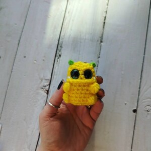 Puede incluir: Una peque&ntilde;a criatura amarilla de crochet con orejas verdes y grandes ojos negros. La criatura est&aacute; sosteniendo sus manos frente a ella.