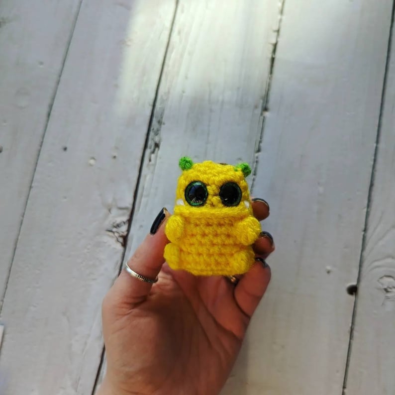 Puede incluir: Una peque&ntilde;a criatura amarilla de crochet con orejas verdes y grandes ojos negros. La criatura est&aacute; sosteniendo sus manos frente a ella.