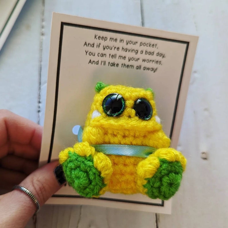 Puede incluir: Un monstruo de crochet amarillo con pies verdes y grandes ojos saltones. El monstruo sostiene una cinta azul y est&aacute; sentado en una tarjeta que dice "Gu&aacute;rdame en tu bolsillo, Y si est&aacute;s teniendo un mal d&iacute;a. Puedes contarme tus preocupaciones, &iexcl;Y yo me las llevar&eacute; todas!"