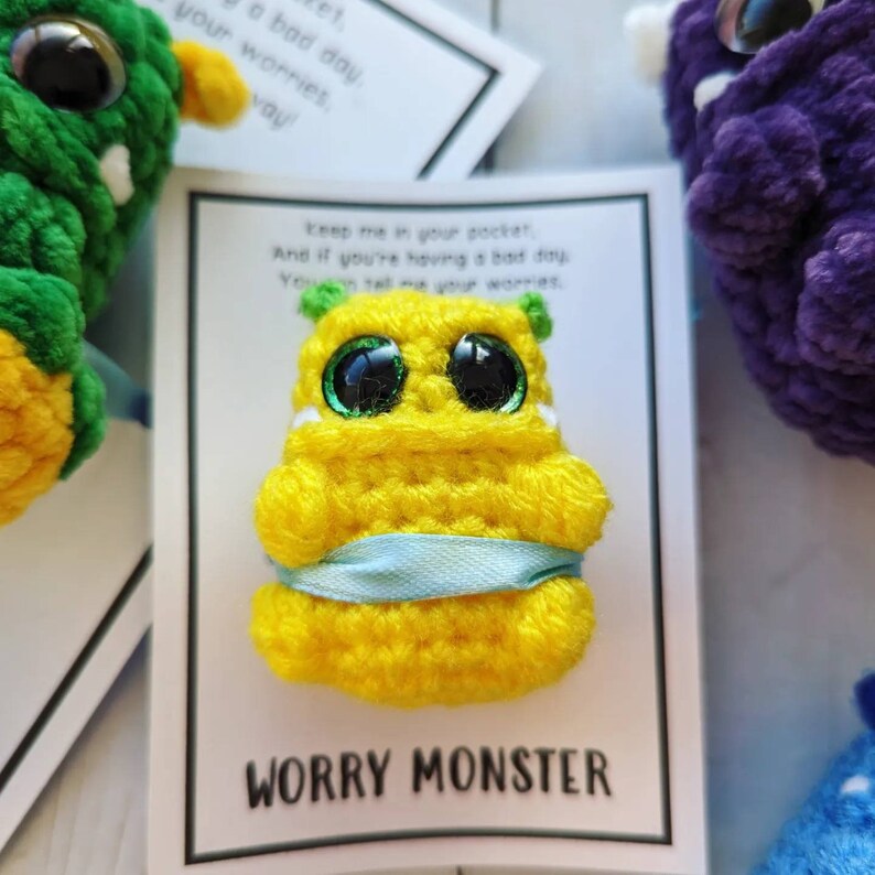 Puede incluir: Monstruo de la preocupaci&oacute;n de color amarillo tejido a crochet con grandes ojos verdes y una cinta azul. El monstruo est&aacute; en una tarjeta blanca con el texto "Gu&aacute;rdame en tu bolsillo. Y si est&aacute;s teniendo un mal d&iacute;a. Puedes contarme tus preocupaciones." y "MONSTRUO DE LA PREOCUPACI&Oacute;N".