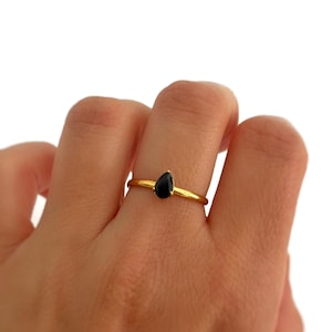 Puede incluir: Un anillo de oro con una piedra preciosa negra en forma de lágrima. El anillo es fino y delicado, con la piedra engastada en un diseño sencillo y elegante. El anillo se muestra en un dedo sobre un fondo blanco.