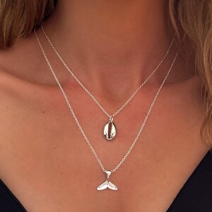 May include: Two silver necklaces with a seashell and a mermaid tail pendant. The necklaces are layered on a person's neck.