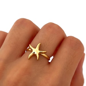 Op de afbeelding: Een goudkleurige ring met een zeesterontwerp. De ring wordt aan een vinger gedragen en toont de gedetailleerde vorm van de zeester. De ring is een sieraad, waarschijnlijk gemaakt van metaal.
