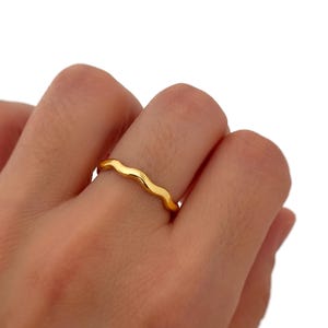 Peut inclure: Une fine bague en or poli présentant un design ondulé ou sinueux continu qui entoure le doigt. L'anneau possède une surface métallique lisse et réfléchissante avec une teinte dorée jaune uniforme sur toute sa structure.