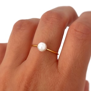 Könnte beinhalten: Ein zarter goldener Ring mit einer einzelnen, runden, weißen Perle. Das schlanke Band des Rings ist goldfarben, und die Perle ist mittig auf dem Band. Der Ring wird an einem Finger getragen.