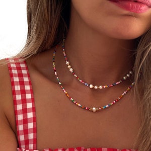 May include: A layered necklace with colorful beads and white pearls. The necklace is worn by a person with long brown hair and a red and white gingham top.