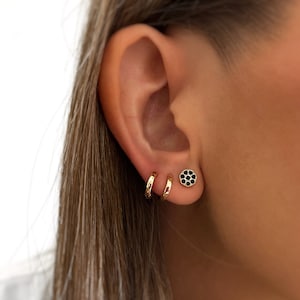 Puede incluir: Aretes de aro de oro con un pequeño pendiente de botón de flor negra en el lóbulo superior de una oreja.