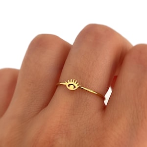 Puede incluir: Un anillo dorado con un diseño de ojo. El anillo tiene una banda delgada y un pequeño dije de ojo estilizado. El dije de ojo tiene una forma curva con un pequeño círculo en el centro y una serie de líneas encima.