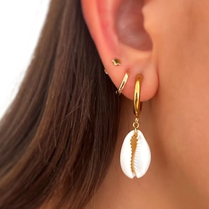 Puede incluir: Pendientes de aro dorados con un colgante de concha blanca. Los pendientes se muestran en una oreja con otros pendientes pequeños dorados. El colgante de concha tiene una superficie texturizada y una forma natural.