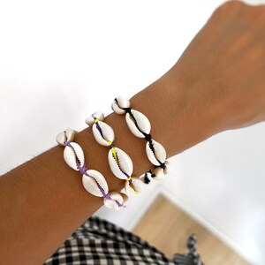 Puede incluir: Tres pulseras de conchas con hilos de diferentes colores. Las pulseras son moradas, amarillas y negras.