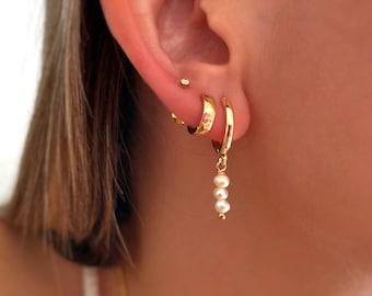 Pode incluir: Brincos de argola de ouro com um pequeno pingente de pérola. As argolas são martelada e têm um acabamento texturizado.