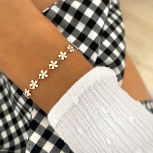 Peut inclure: Un bracelet en chaîne dorée avec des breloques en forme de marguerite blanche. Le bracelet est porté au poignet.