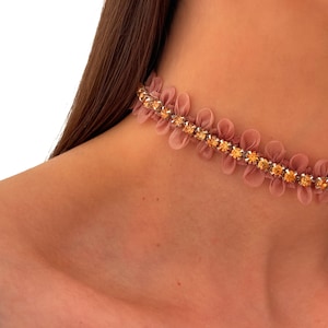 Może przedstawiać: Naszyjnik typu choker z centralnym rzędem błyszczących bursztynowych cyrkonii osadzonych w srebrnym metalowym łańcuszku, otoczony delikatnym, marszczonym wykończeniem z pudrowo-różowej tkaniny, tworzącym miękką, warstwową teksturę wokół szyi.