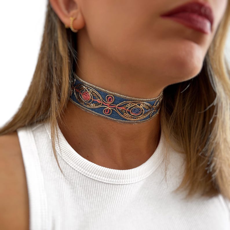 May include: A blue choker necklace with gold and pink embroidery. The choker is made of a fabric ribbon with a geometric pattern.