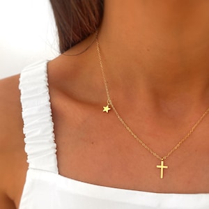 Puede incluir: Un collar dorado con dos colgantes: una pequeña estrella y una cruz. La fina cadena se muestra sobre una parte superior blanca. La estrella cuelga cerca de la clavícula, la cruz más abajo.