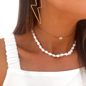 May include: A layered necklace set featuring a white shell necklace and a gold chain necklace with a small daisy pendant. A gold lightning bolt earring is visible in the background. The jewelry is worn over a white top.