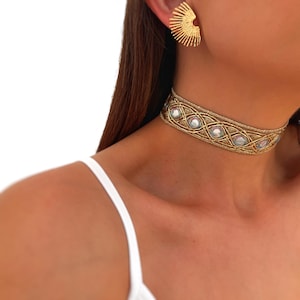 May include: A gold-toned choker necklace with a woven design and iridescent embellishments. The necklace is paired with matching gold fan-shaped earrings. The jewelry is set against a white background.