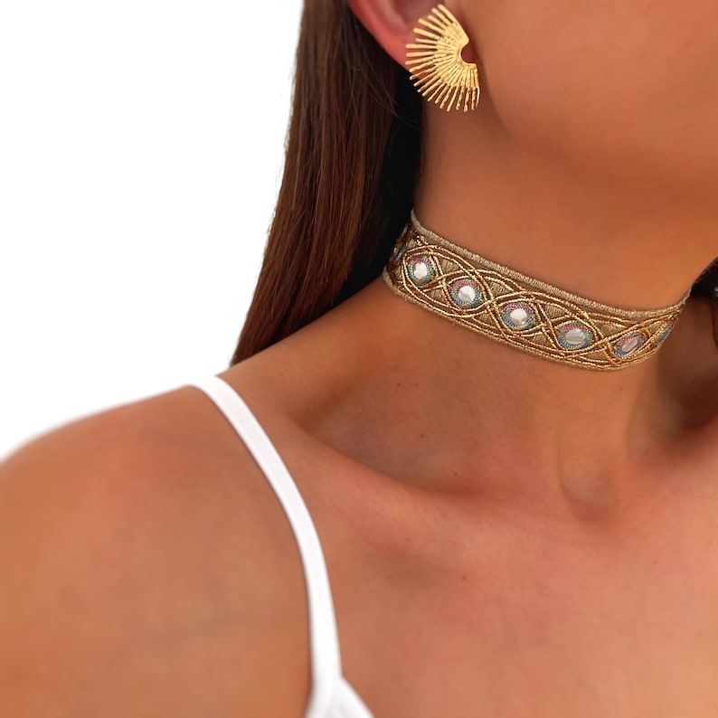 May include: A gold-toned choker necklace with a woven design and iridescent embellishments. The necklace is paired with matching gold fan-shaped earrings. The jewelry is set against a white background.