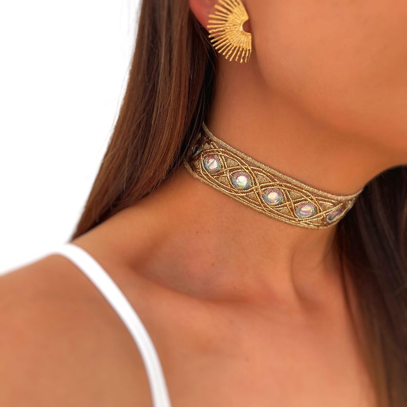 May include: Gold-toned choker necklace with iridescent stones and intricate woven design. The choker is worn around the neck and is paired with a gold sunburst earring. The jewelry is set against a white background.