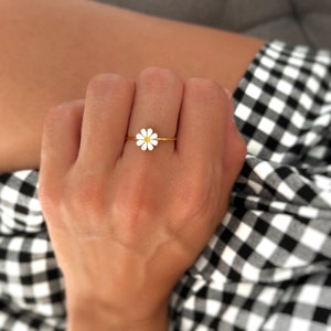 May include: A gold ring featuring a white daisy flower design. The ring is worn on a finger.