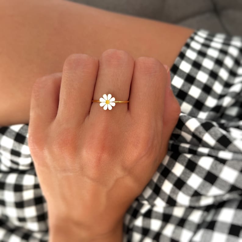 May include: A gold ring featuring a white daisy flower design. The ring is worn on a finger.