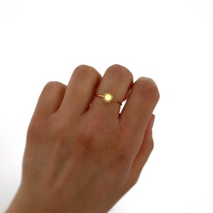 May include: A gold ring with a small sun design. The ring is on a person's finger.