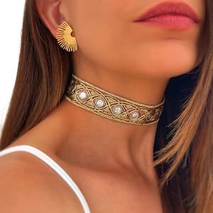 May include: A gold-colored choker necklace with iridescent stones and a matching gold earring. The choker features an intricate woven design with oval-shaped iridescent stones. The earring is a half-circle shape with vertical lines.